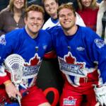Two hockey players in blue jerseys pose and smile for the camera near the rink boards, fans behind them celebrate.