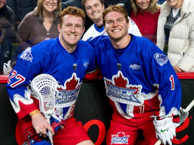 Two hockey players in blue jerseys pose and smile for the camera near the rink boards, fans behind them celebrate.