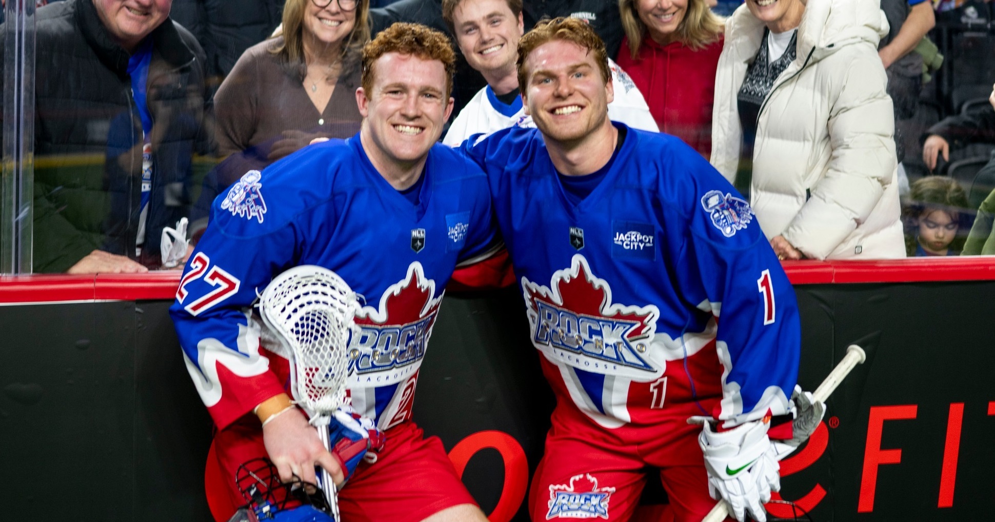 Two hockey players in blue jerseys pose and smile for the camera near the rink boards, fans behind them celebrate.