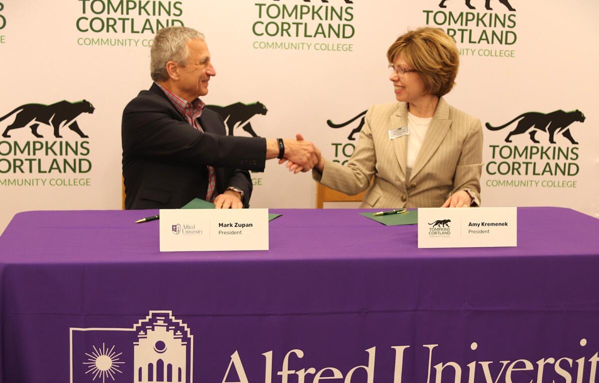 Two professionals shake hands across a purple-clothed table at a college event, with Tompkins Cortland Community College branding in the background.