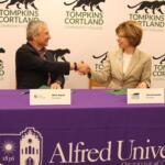 Two professionals shake hands across a purple-clothed table at a college event, with Tompkins Cortland Community College branding in the background.
