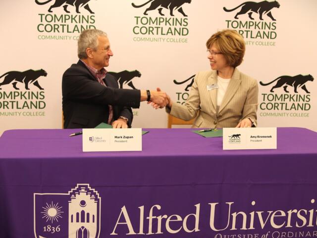 Two professionals shake hands across a purple-clothed table at a college event, with Tompkins Cortland Community College branding in the background.