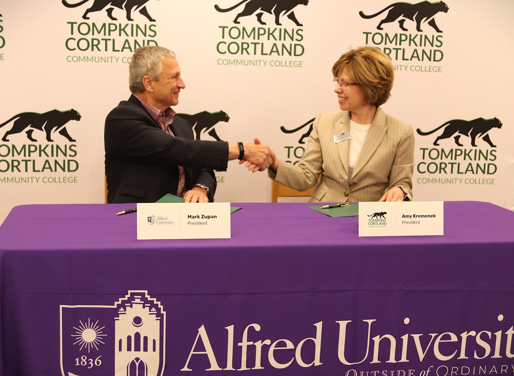 Two professionals shake hands across a purple-clothed table at a college event, with Tompkins Cortland Community College branding in the background.