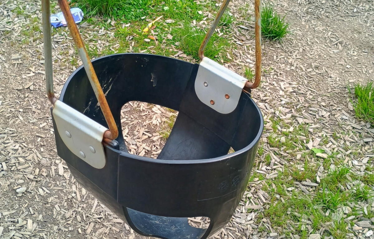 Black plastic bucket swing hanging by rusted metal chains in an outdoor playground on wood chip ground.