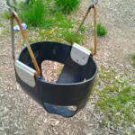 Black plastic bucket swing hanging by rusted metal chains in an outdoor playground on wood chip ground.