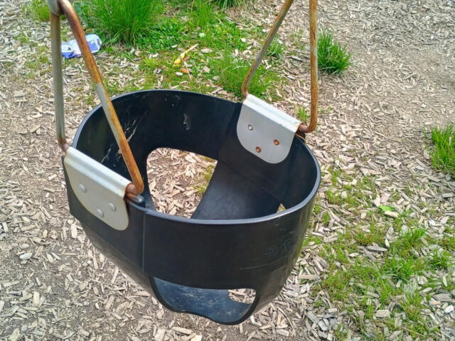 Black plastic bucket swing hanging by rusted metal chains in an outdoor playground on wood chip ground.