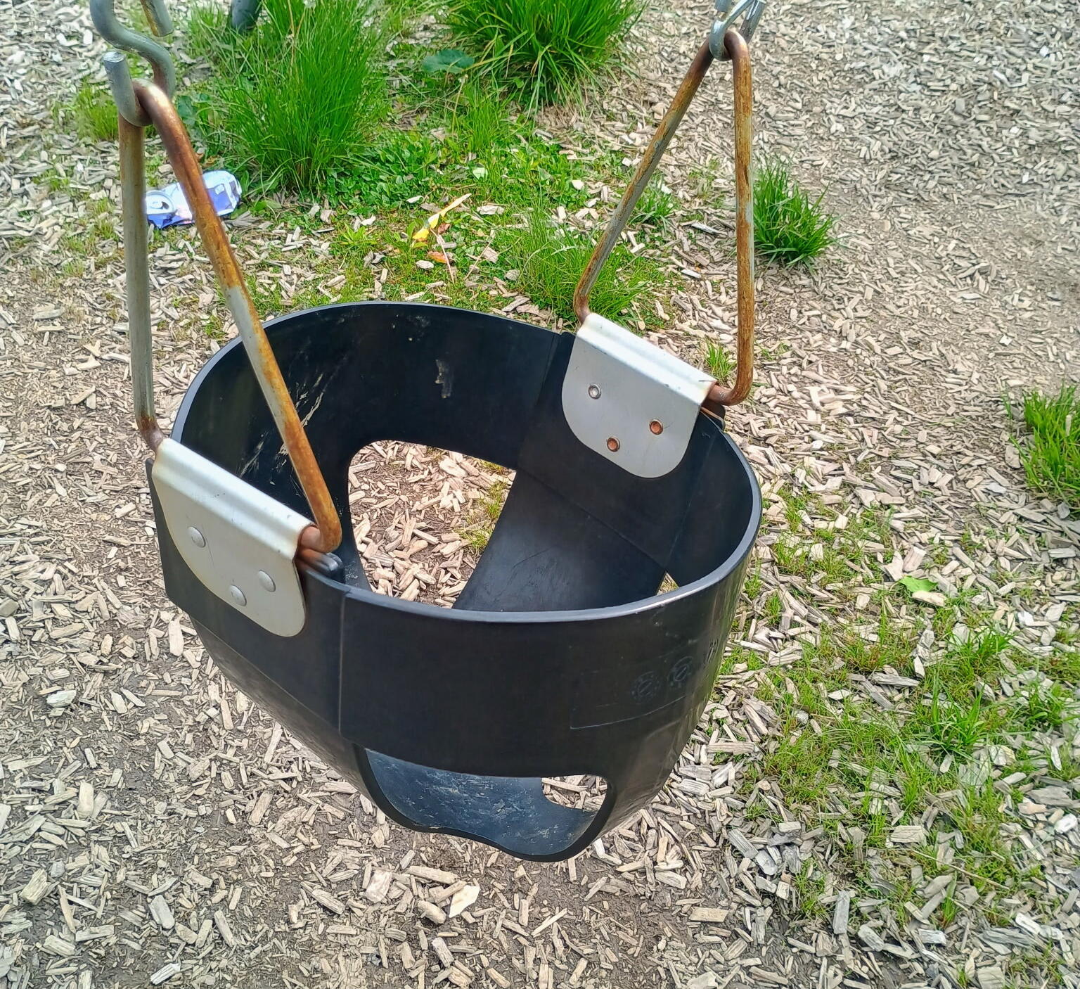 Black plastic bucket swing hanging by rusted metal chains in an outdoor playground on wood chip ground.