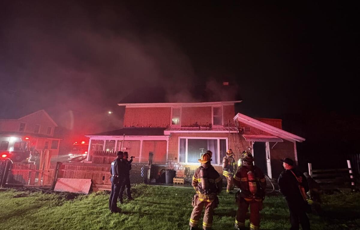 Nighttime scene of a two-story house with smoke rising from the roof as firefighters in gear work outside, red emergency lights from a fire truck nearby.