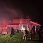 Nighttime scene of a two-story house with smoke rising from the roof as firefighters in gear work outside, red emergency lights from a fire truck nearby.