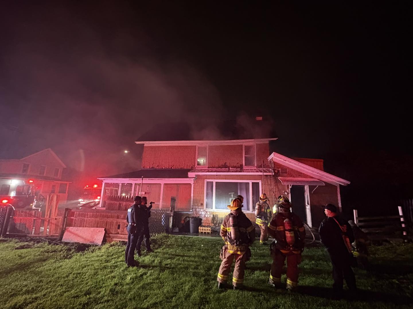 Nighttime scene of a two-story house with smoke rising from the roof as firefighters in gear work outside, red emergency lights from a fire truck nearby.