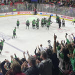 Green-uniform hockey players celebrate on the ice as a cheering crowd watches from the stands.