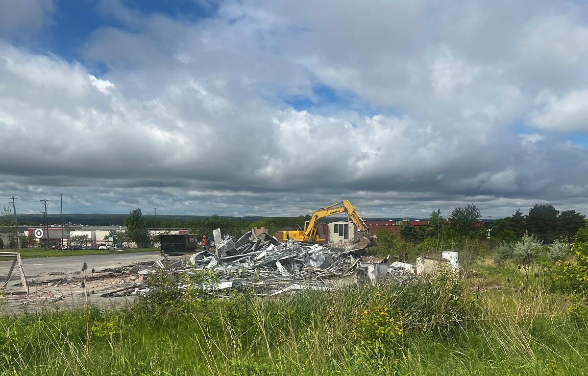 Yellow excavator on a demolition site tossing metal debris into a pile, with tall grass in the foreground and a cloudy sky above.