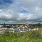 Yellow excavator on a demolition site tossing metal debris into a pile, with tall grass in the foreground and a cloudy sky above.