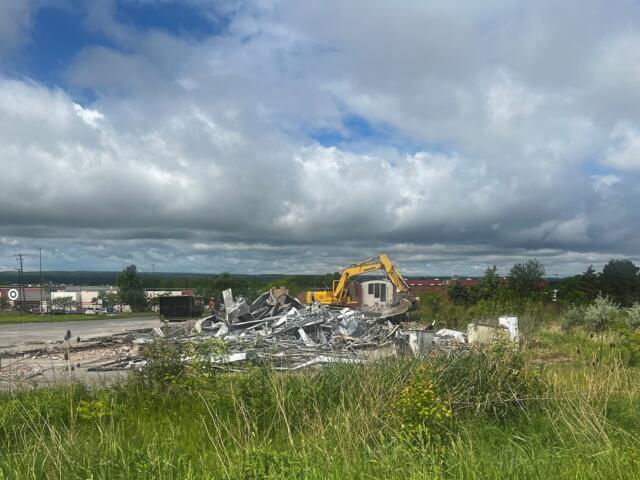 Yellow excavator on a demolition site tossing metal debris into a pile, with tall grass in the foreground and a cloudy sky above.