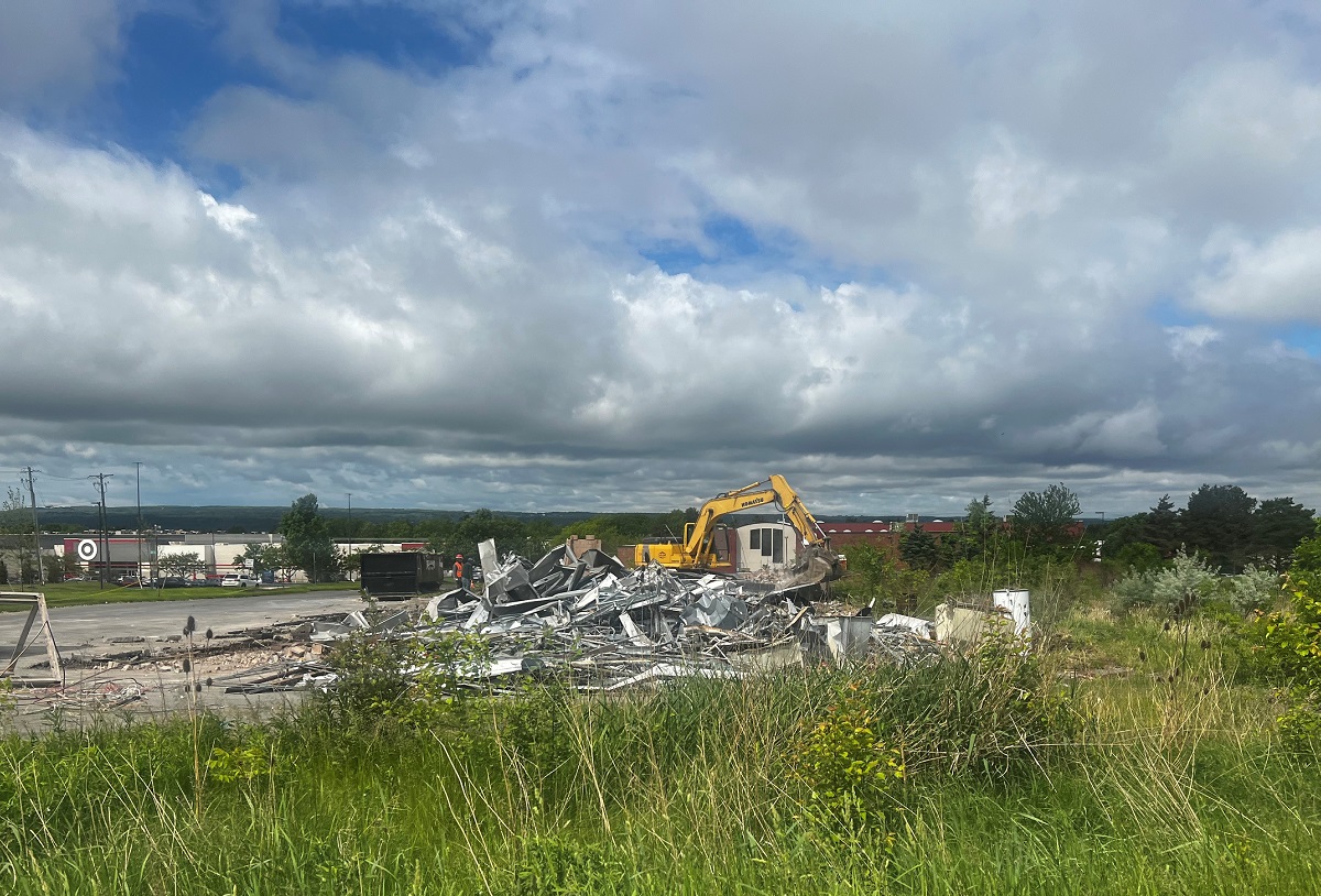 Yellow excavator on a demolition site tossing metal debris into a pile, with tall grass in the foreground and a cloudy sky above.