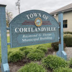 Blue signboard for the Town of Cortlandville with its seal and 'Raymond G. Thorpe Municipal Building' text in front of shrubs on a cloudy day