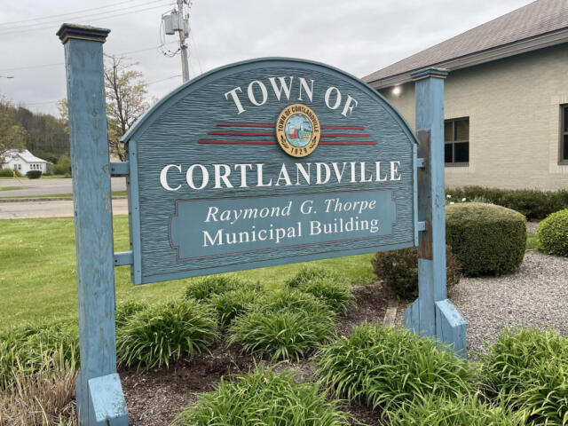 Blue signboard for the Town of Cortlandville with its seal and 'Raymond G. Thorpe Municipal Building' text in front of shrubs on a cloudy day