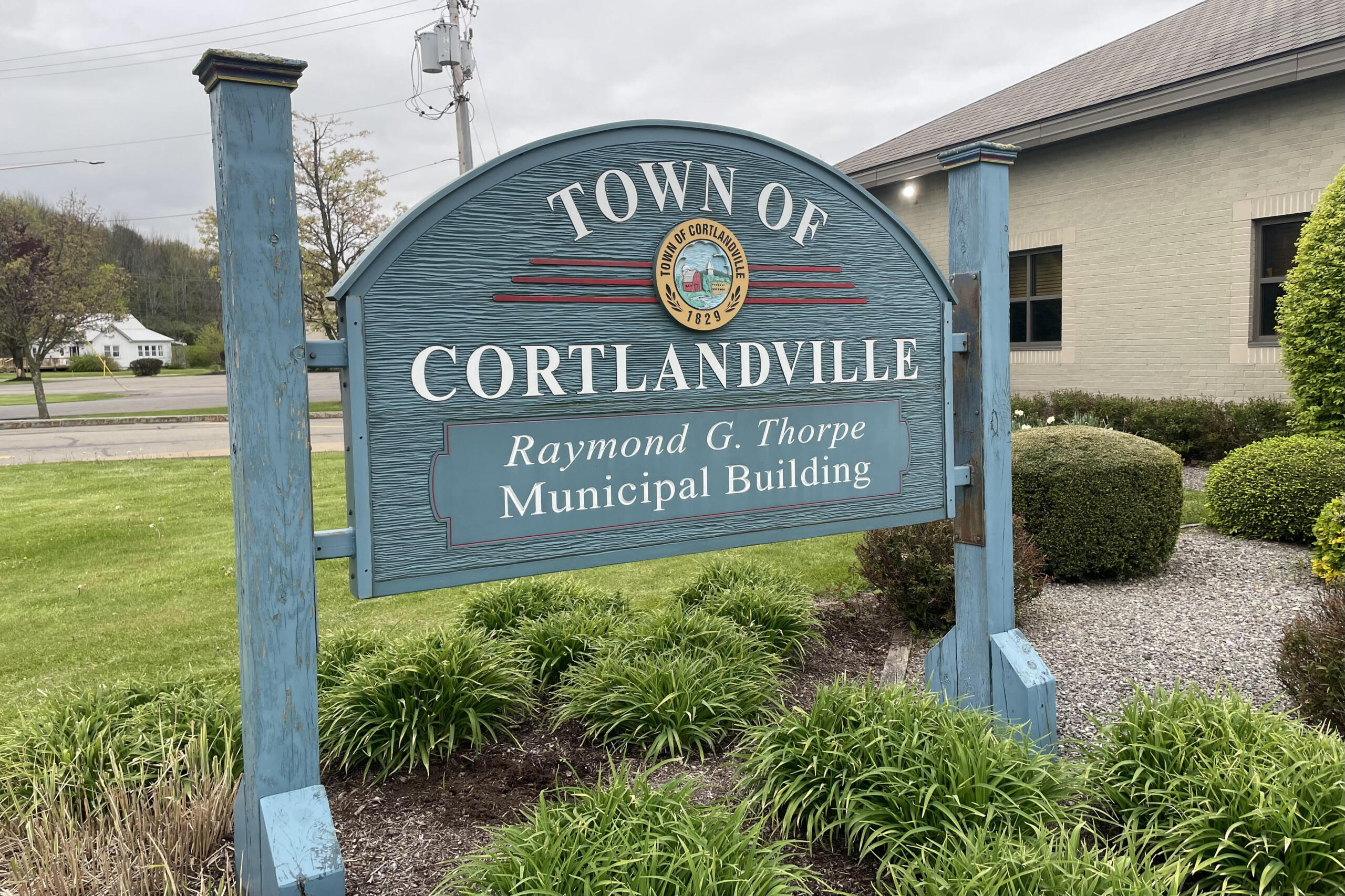 Blue signboard for the Town of Cortlandville with its seal and 'Raymond G. Thorpe Municipal Building' text in front of shrubs on a cloudy day