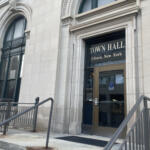 Front of Town Hall Ithaca, New York, showing a stone facade, arched windows, and a glass door with beige frame behind metal railings.
