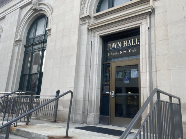 Front of Town Hall Ithaca, New York, showing a stone facade, arched windows, and a glass door with beige frame behind metal railings.