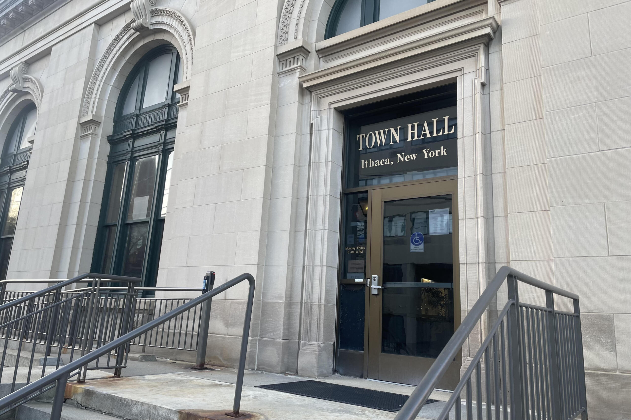 Front of Town Hall Ithaca, New York, showing a stone facade, arched windows, and a glass door with beige frame behind metal railings.