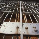 Close-up of a metal jail gate with vertical bars and a riveted control panel, showing rust and a latch area behind the bars.