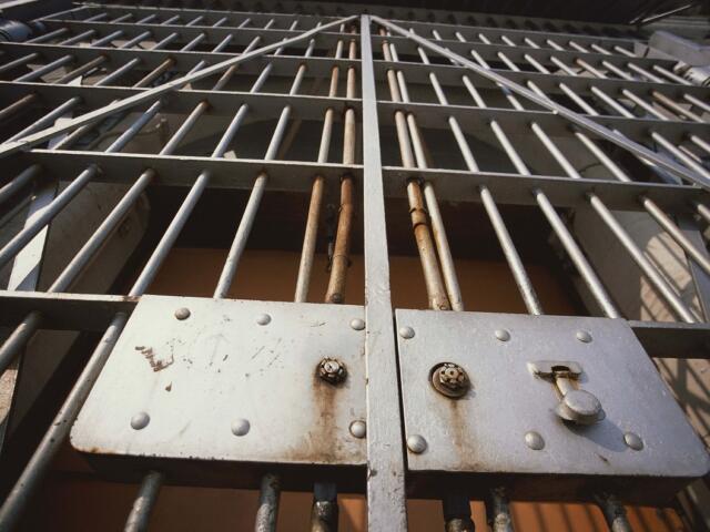 Close-up of a metal jail gate with vertical bars and a riveted control panel, showing rust and a latch area behind the bars.