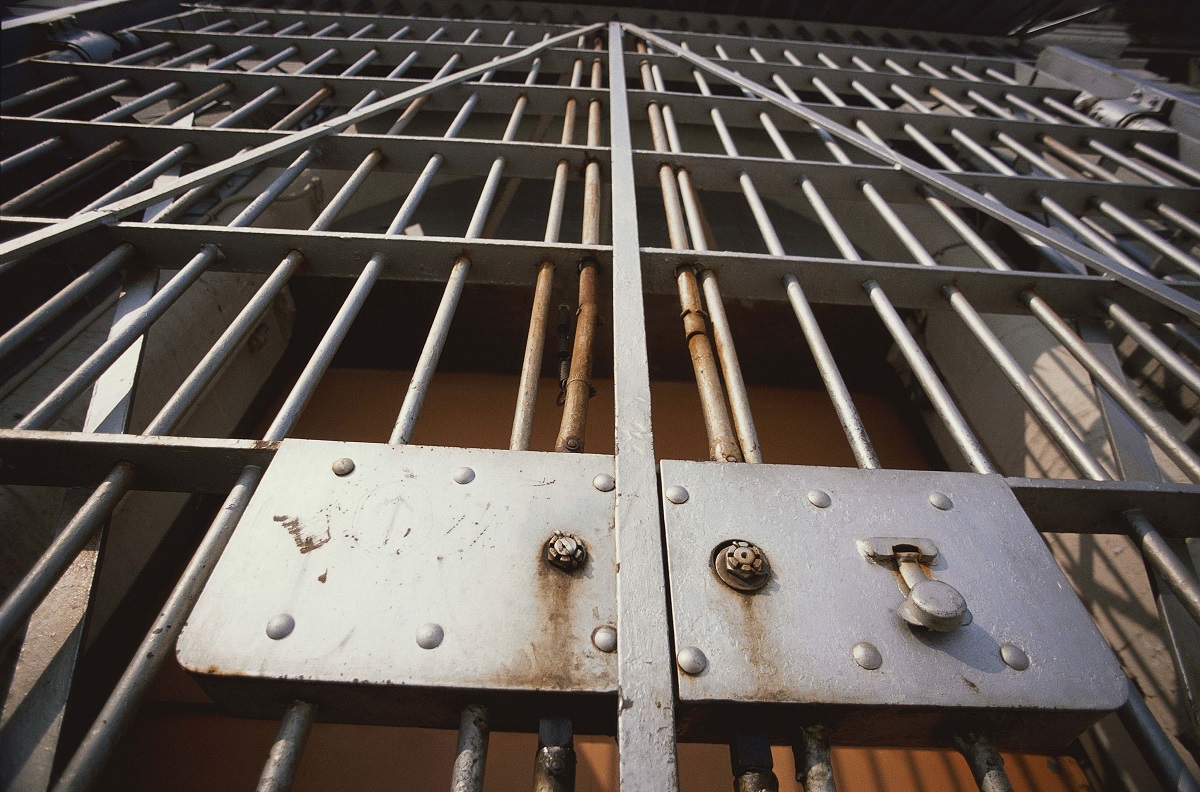 Close-up of a metal jail gate with vertical bars and a riveted control panel, showing rust and a latch area behind the bars.
