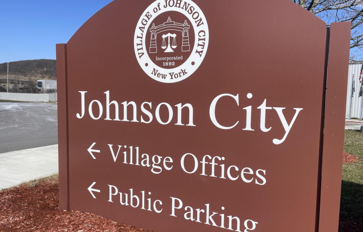 Brown Johnson City sign featuring a white circular city seal and large 'Johnson City' text; indicates Village Offices directions in an outdoor setting.