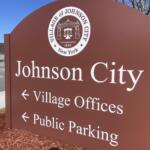 Brown Johnson City sign featuring a white circular city seal and large 'Johnson City' text; indicates Village Offices directions in an outdoor setting.