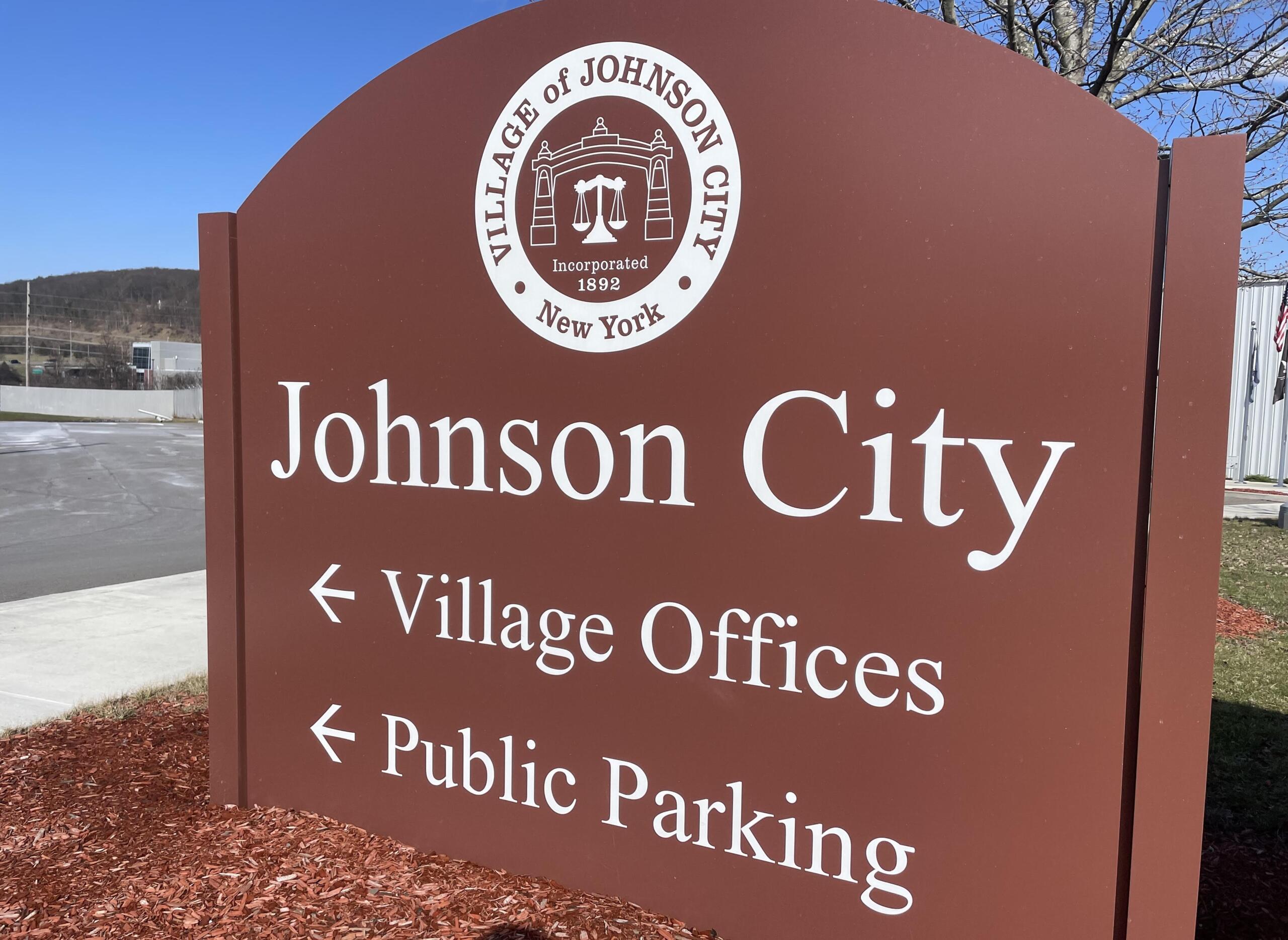 Brown Johnson City sign featuring a white circular city seal and large 'Johnson City' text; indicates Village Offices directions in an outdoor setting.