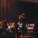 Conductor leading a full orchestra on stage during a performance and musicians playing violins nearby.