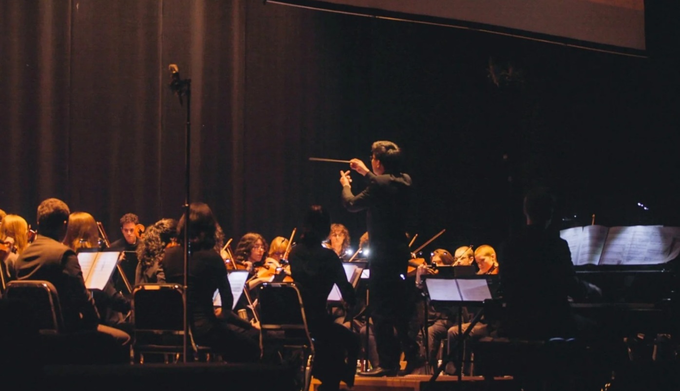 Conductor leading a full orchestra on stage during a performance and musicians playing violins nearby.