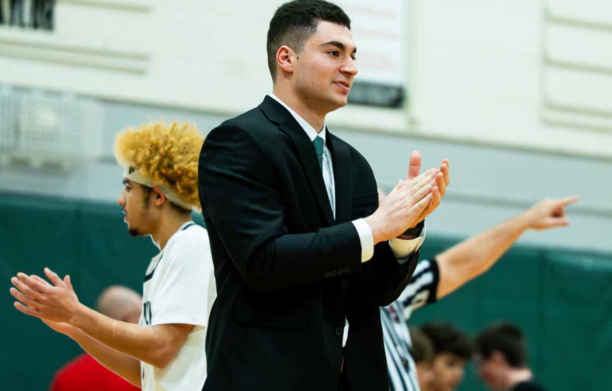 Young man in a black suit clapping at a gym with players in white uniforms in the background.