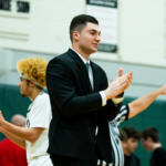 Young man in a black suit clapping at a gym with players in white uniforms in the background.