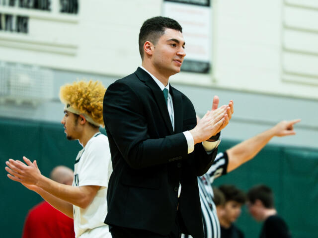 Young man in a black suit clapping at a gym with players in white uniforms in the background.