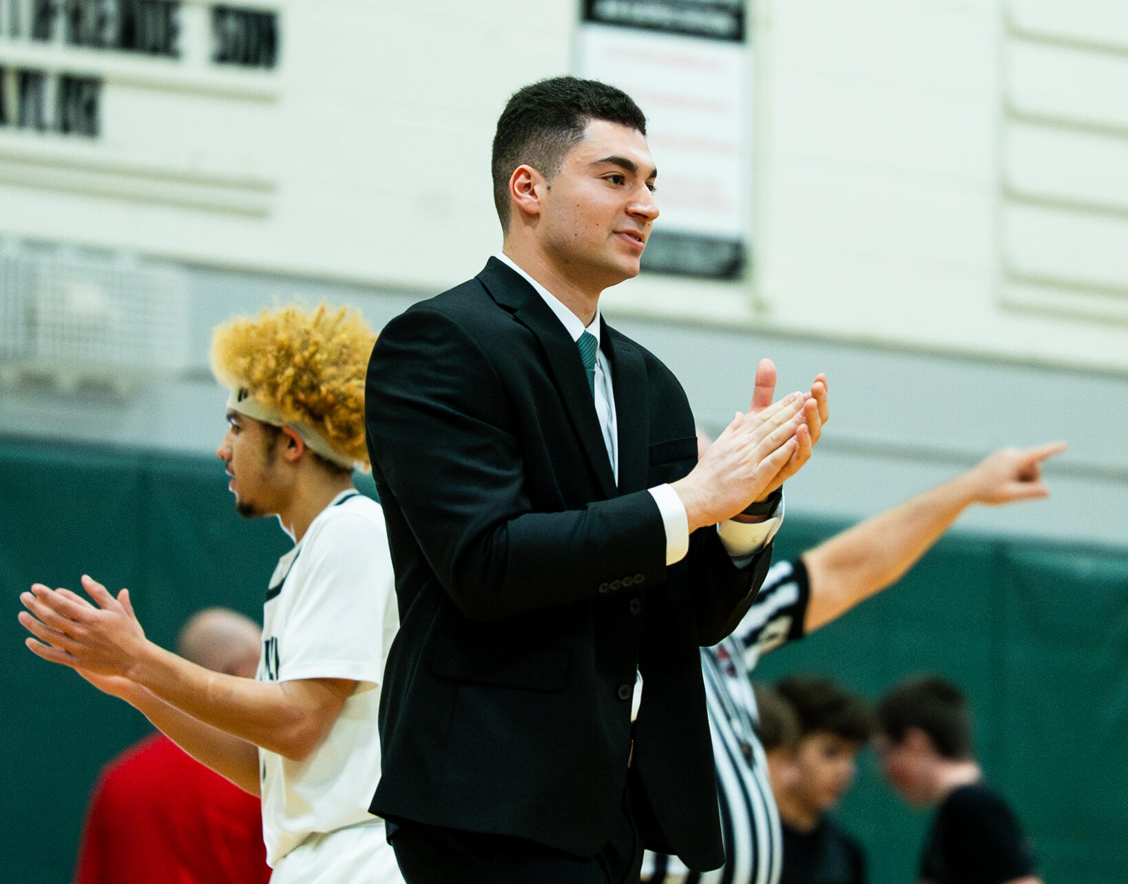 Young man in a black suit clapping at a gym with players in white uniforms in the background.