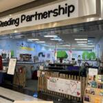 Family Reading Partnership storefront in a mall with a small fenced reading area, bookshelves, and tree murals on blue walls.