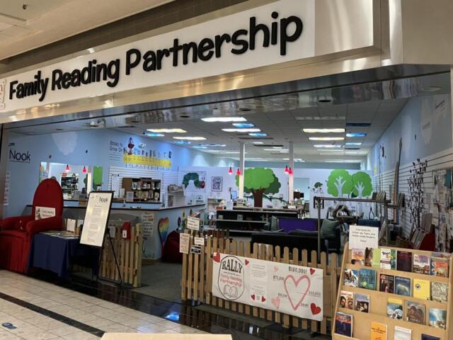 Family Reading Partnership storefront in a mall with a small fenced reading area, bookshelves, and tree murals on blue walls.