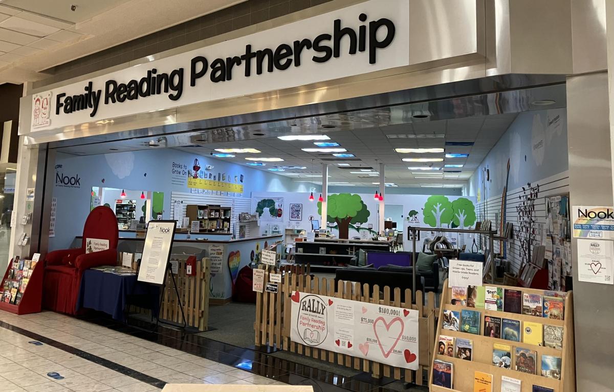 Family Reading Partnership storefront in a mall with a small fenced reading area, bookshelves, and tree murals on blue walls.