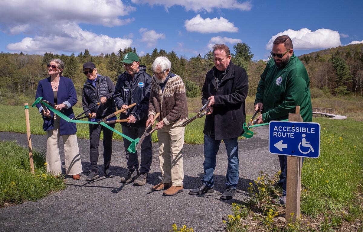 Seven adults take part in a ribbon-cutting ceremony for an accessible-path project along a trail, with a blue Accessible Route sign nearby.