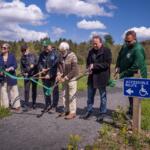 Seven adults take part in a ribbon-cutting ceremony for an accessible-path project along a trail, with a blue Accessible Route sign nearby.