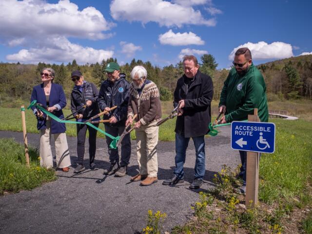 Seven adults take part in a ribbon-cutting ceremony for an accessible-path project along a trail, with a blue Accessible Route sign nearby.