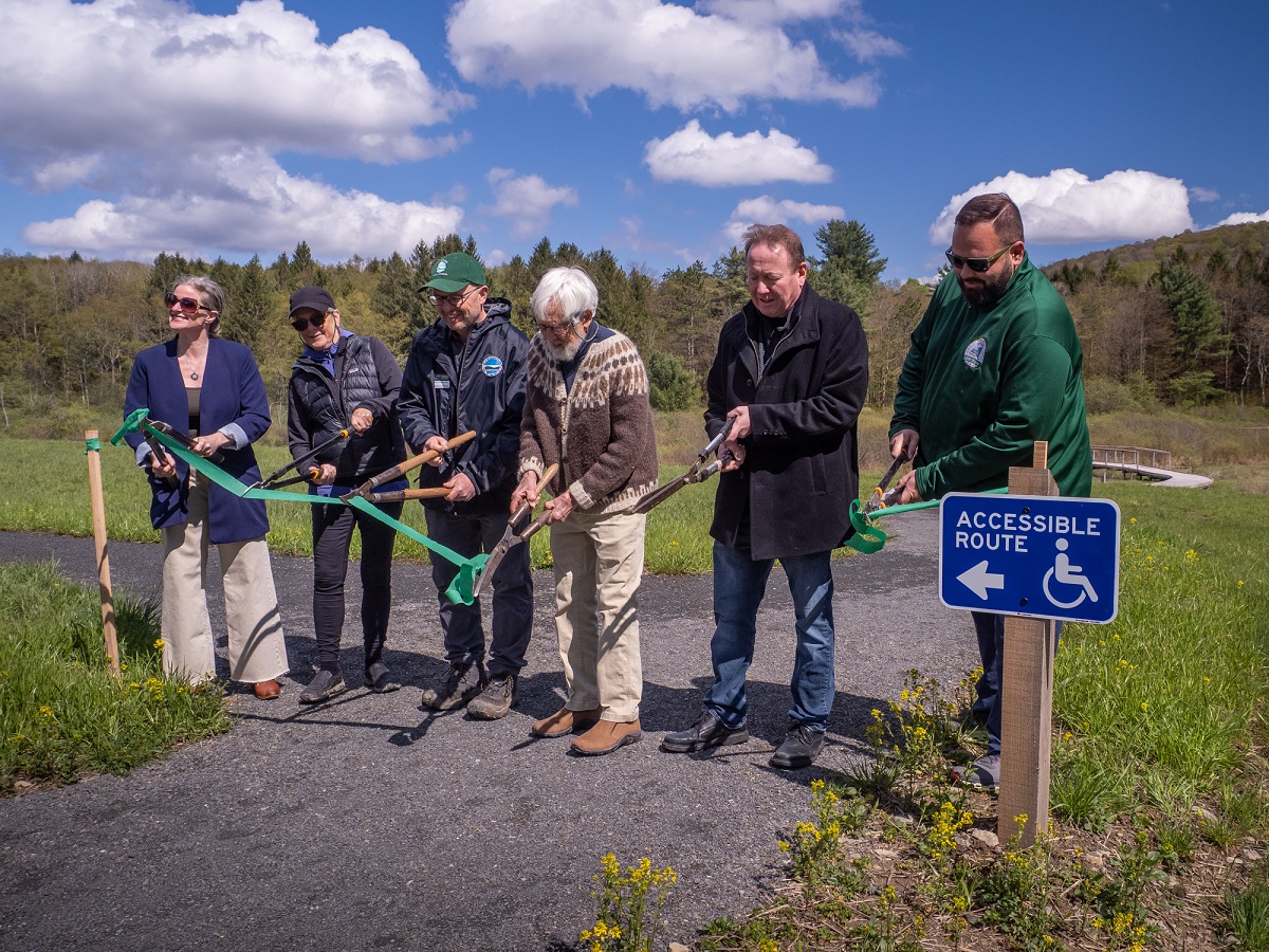 Seven adults take part in a ribbon-cutting ceremony for an accessible-path project along a trail, with a blue Accessible Route sign nearby.