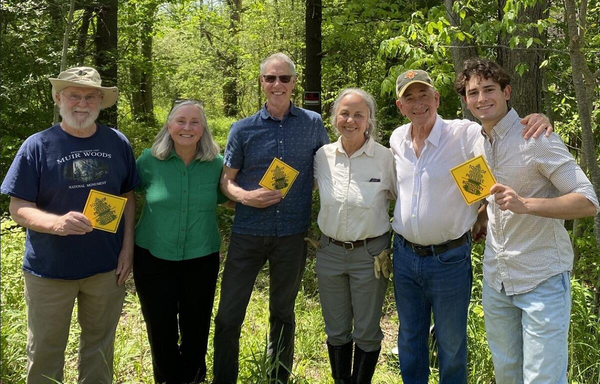 Six adults stand in a sunlit forest, each holding a yellow diamond sign and smiling at the camera.