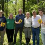 Six adults stand in a sunlit forest, each holding a yellow diamond sign and smiling at the camera.