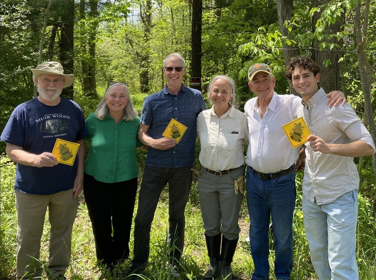 Six adults stand in a sunlit forest, each holding a yellow diamond sign and smiling at the camera.