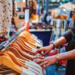 Person browsing a clothing rack in a store, reaching for a garment on wooden hangers.