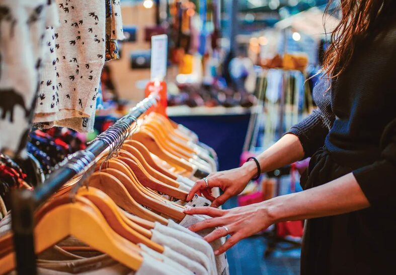 Person browsing a clothing rack in a store, reaching for a garment on wooden hangers.