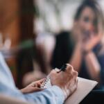 Therapist taking notes during a counseling session, client visible in the background blurred.